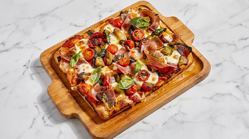 Rectangular pizza with prosciutto, cherry tomatoes, mozzarella, basil, and roasted vegetables on a wooden board.