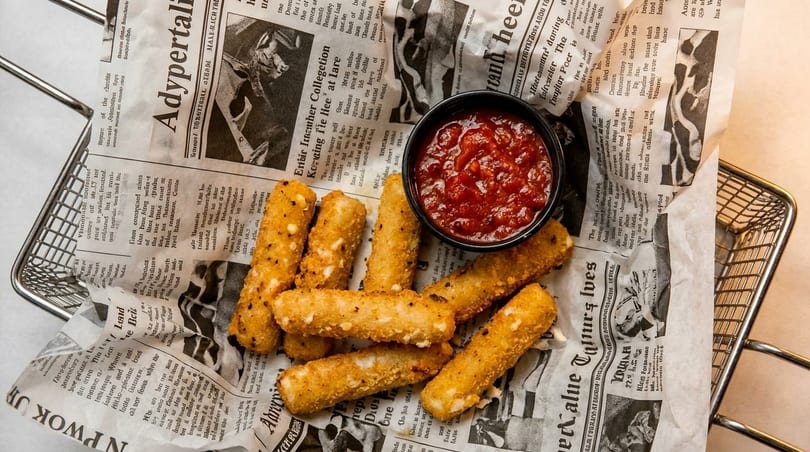 Crispy golden mozzarella sticks and a small bowl of marinara sauce in a metal basket lined with newspaper.