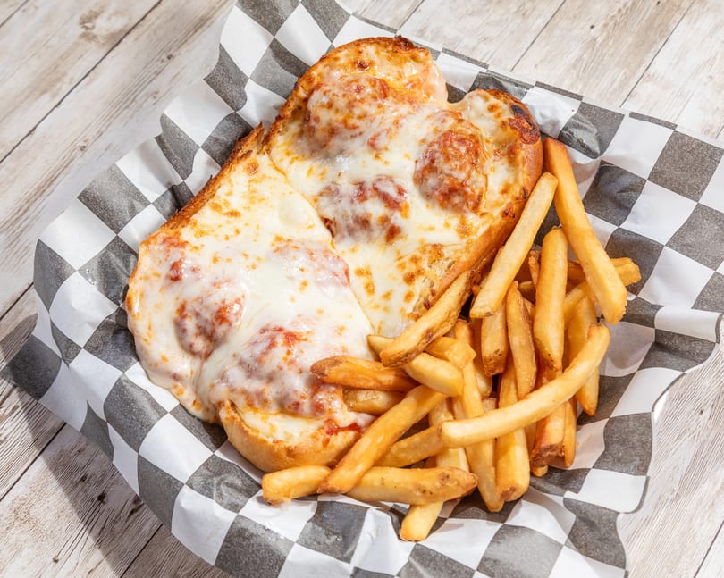 Meatball sub with melted cheese and a side of golden french fries in a checkered paper-lined basket.