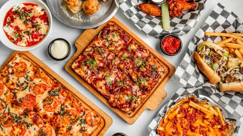 A flat lay of various dishes including two square pizzas, a Philly cheesesteak, and loaded fries.