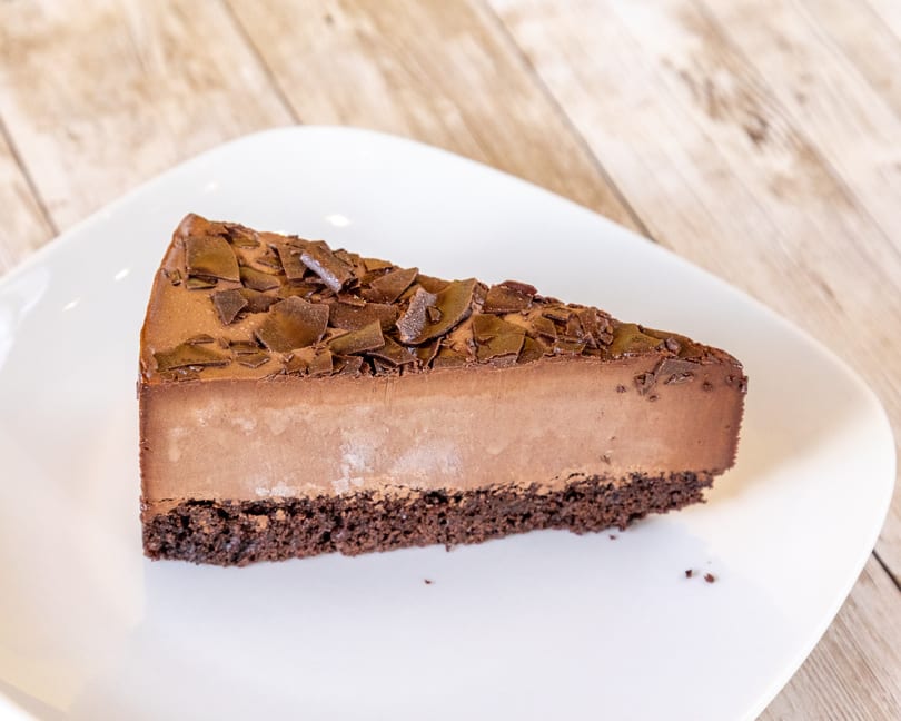 A slice of rich chocolate cheesecake with chocolate shavings on top, on a white plate.
