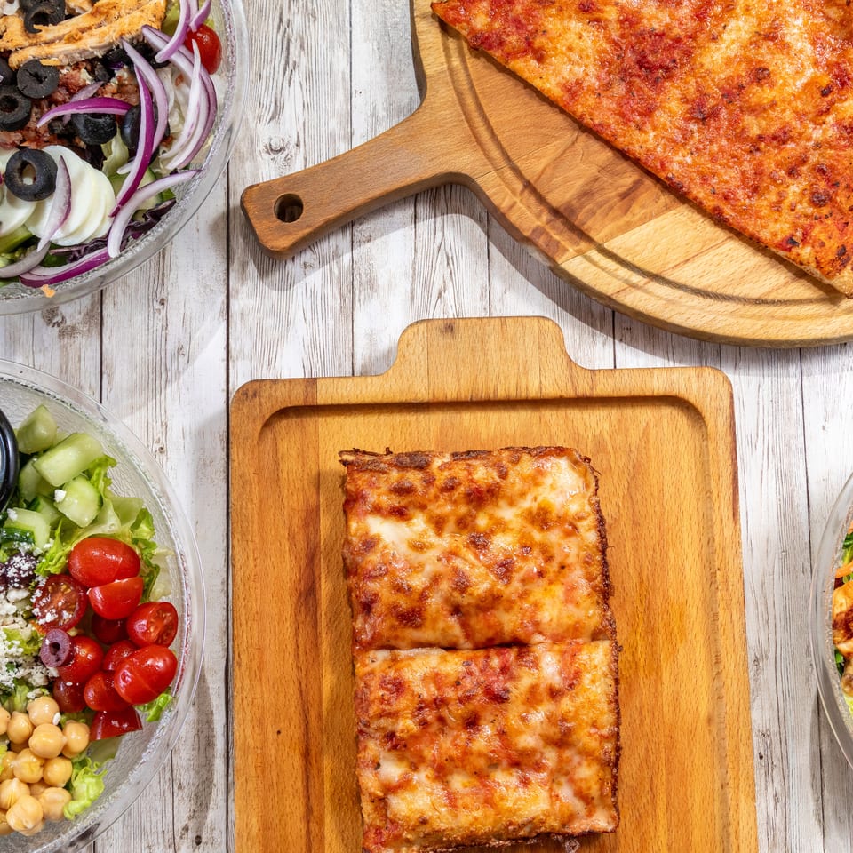 Overhead view of three fresh salads, a cheese pizza slice, and two square pizza slices on wooden boards.
