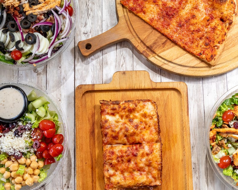 Overhead view of three fresh salads, a cheese pizza slice, and two square pizza slices on wooden boards.