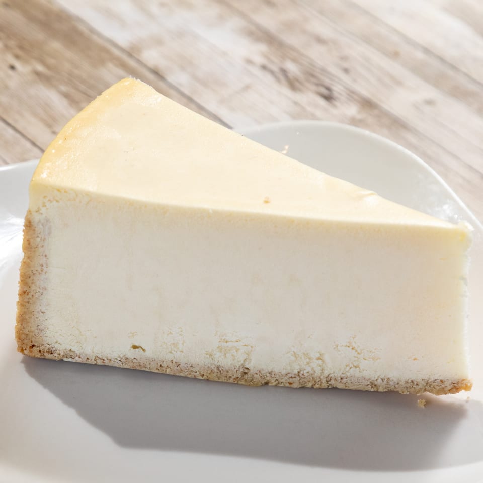 Slice of classic cheesecake with a graham cracker crust on a white plate.