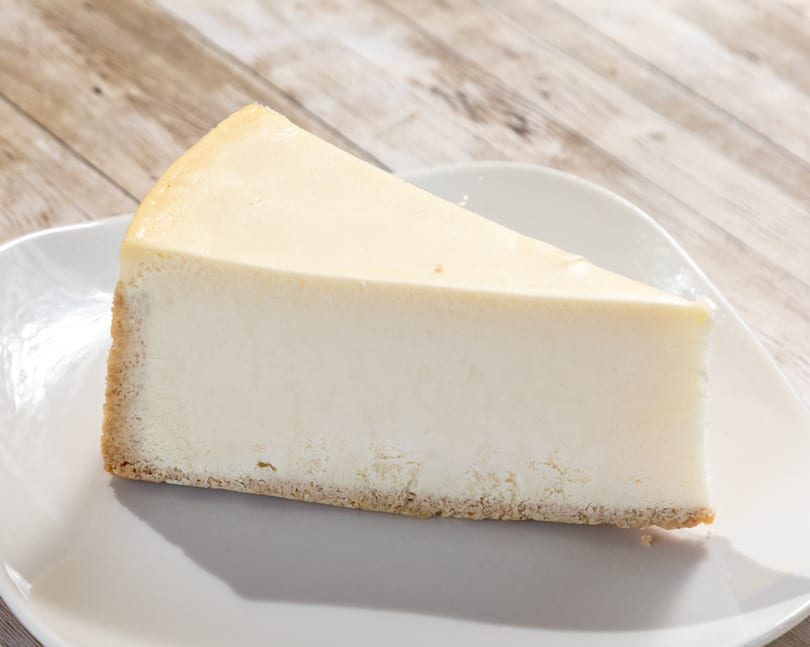 Slice of classic cheesecake with a graham cracker crust on a white plate.